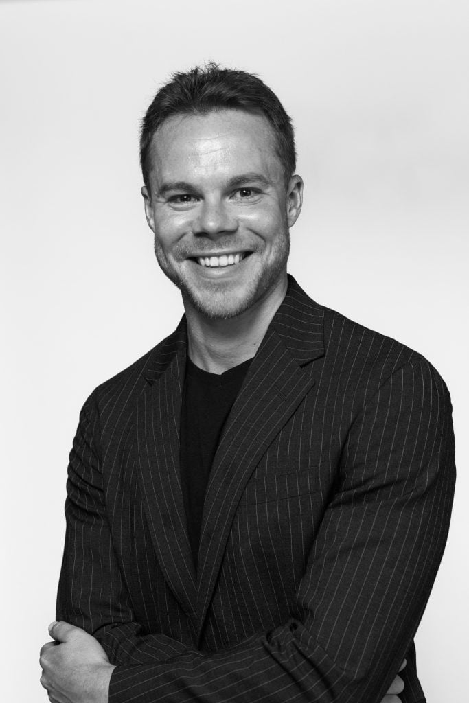 Black-and-white half portrait of Edward Warhburton, owner of art advisory and dealership Princedale Modern. Wearing a black crew shirt and blazer smiling at the camera.
