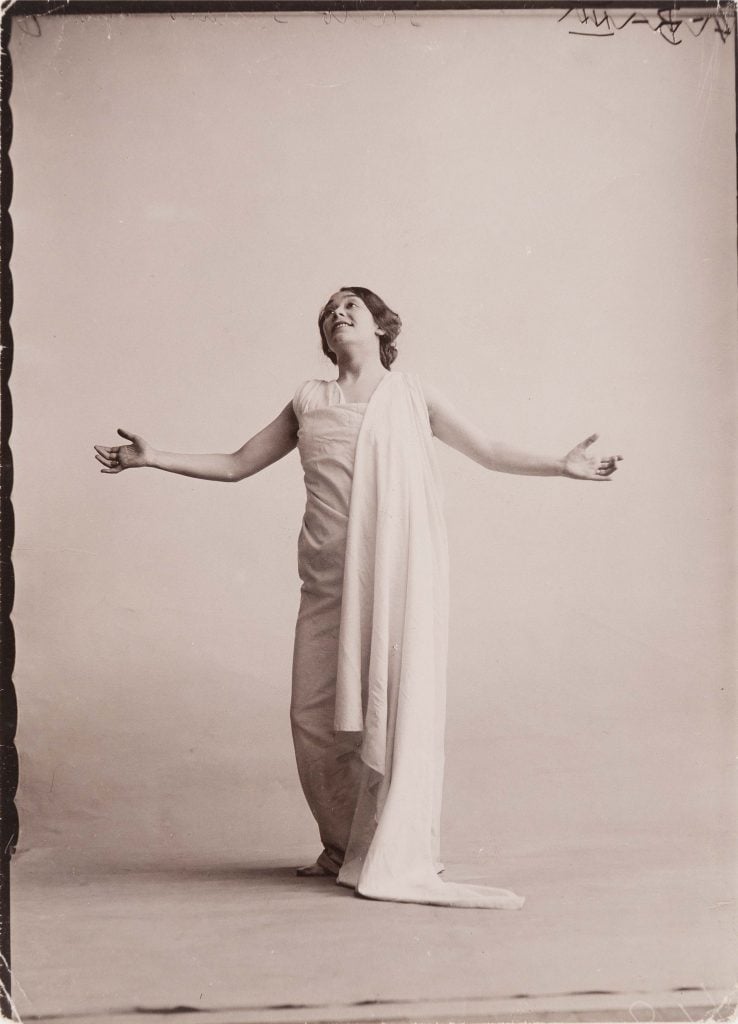 A black and white image chosen by pascal rousseau of an individual in white drapery and costume with arms outspread in a dance move.