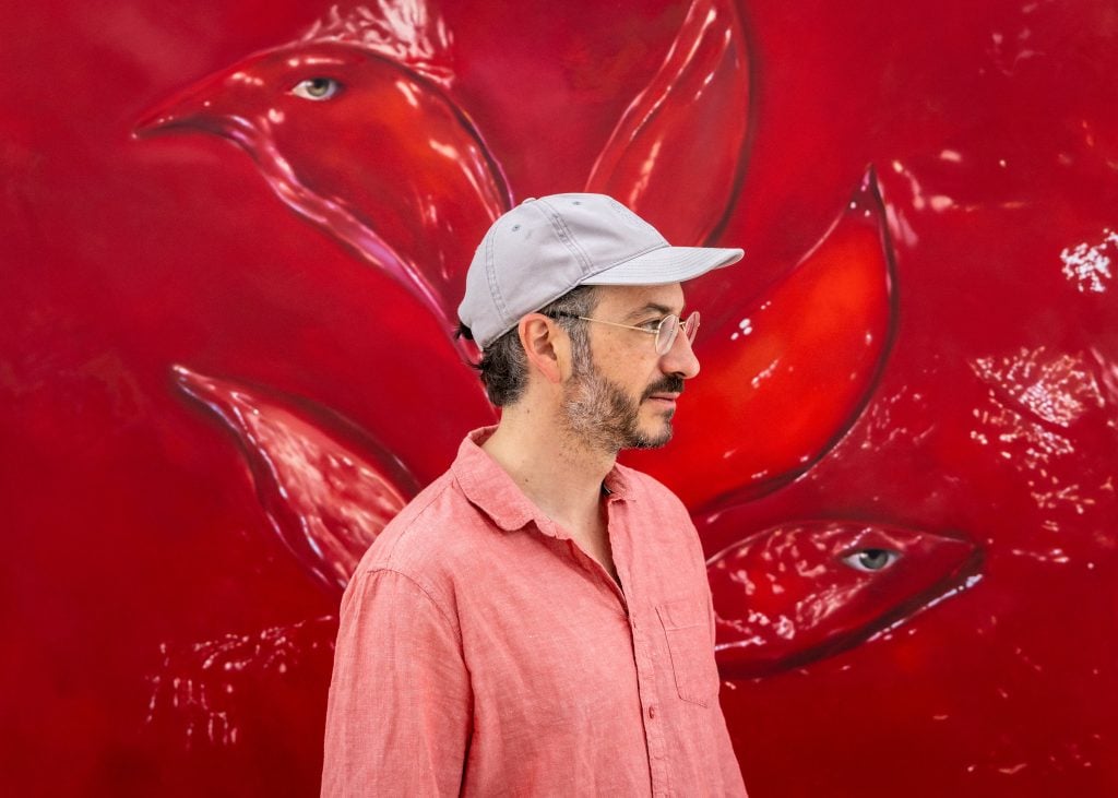 a profile photograph of a man in a light gray ball cap. he is standing in front of a big red painting of a bird. the man is wearing a red polo shirt. he's wearing a pair of glasses and has a beard and mustache,