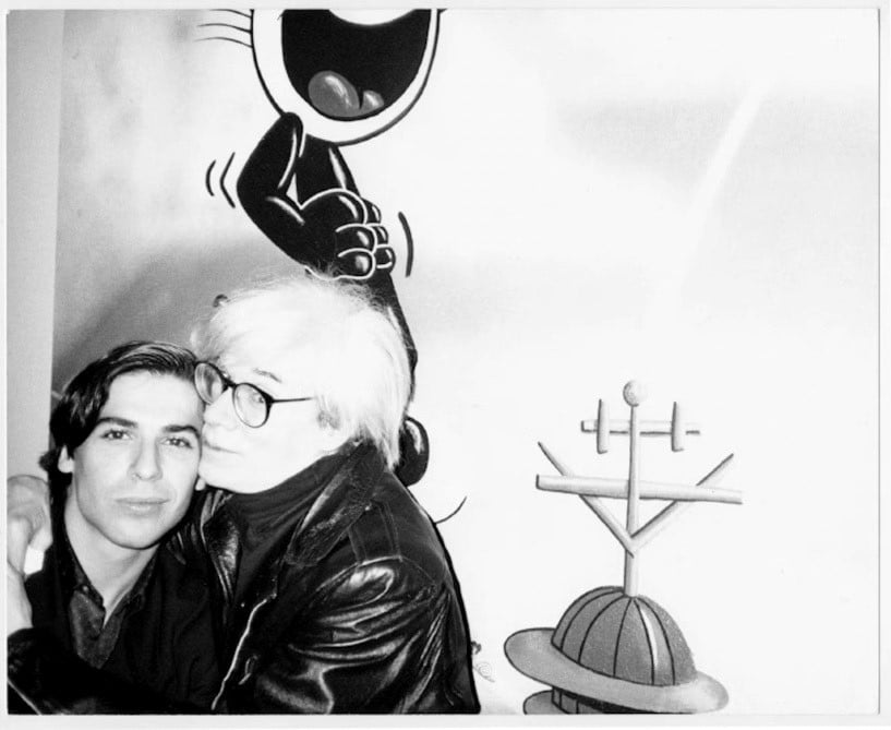 a black and white photo of two men embracing. one of the men is andy warhol in his signature fright wig and glasses, the other is the artist george condo