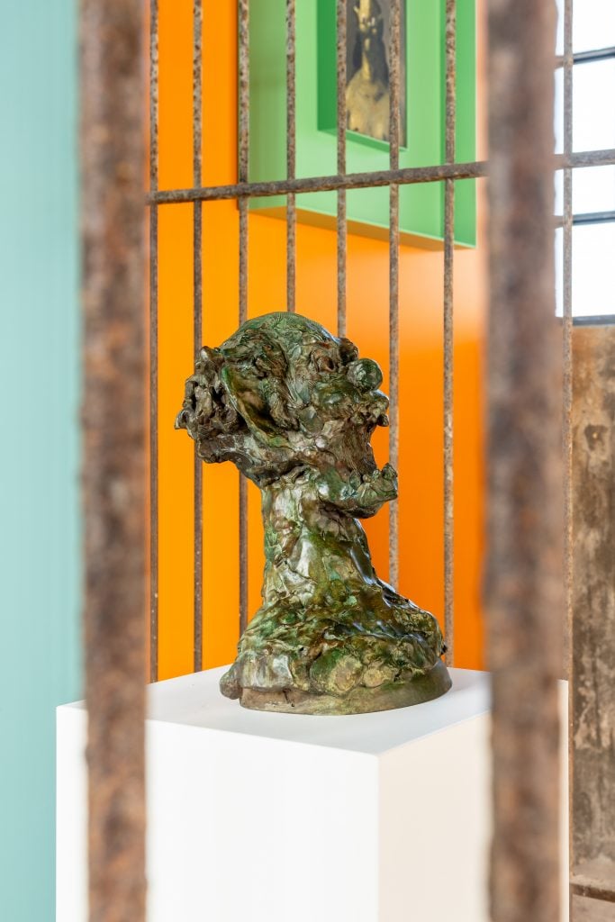 a distorted bronze head sculpture in a cage against an orange wall