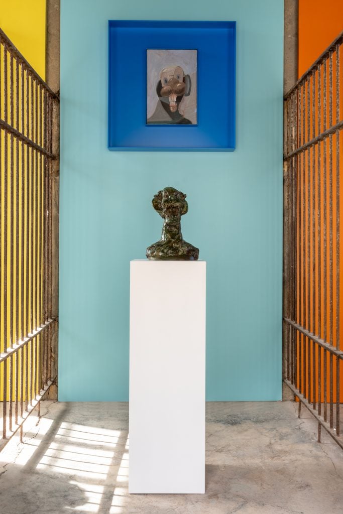 a distorted bronze head sculpture in a cage against a blue wall