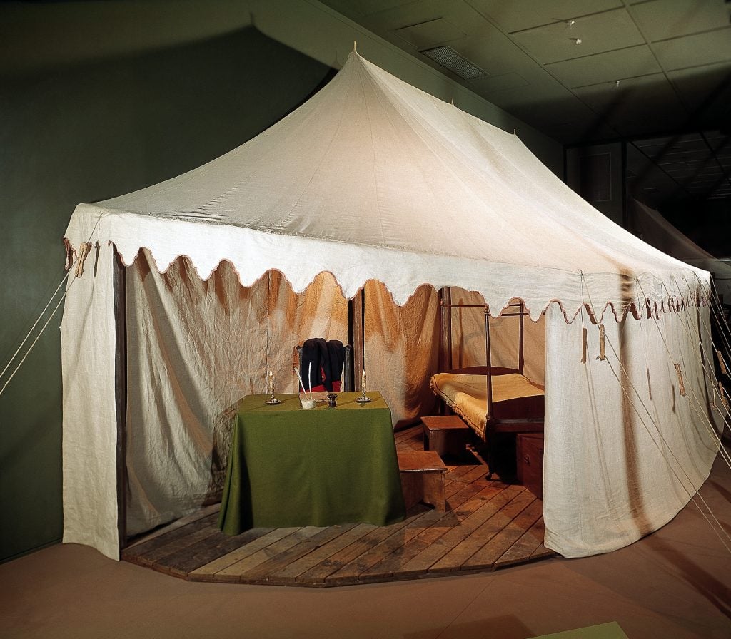 A white linen tent with scallop red-trimmed edges used by George Washington during the Revolutionary War. The tent is erected in a darkened room, with a four-post bed and desk covered with an olive green tablecloth inside.