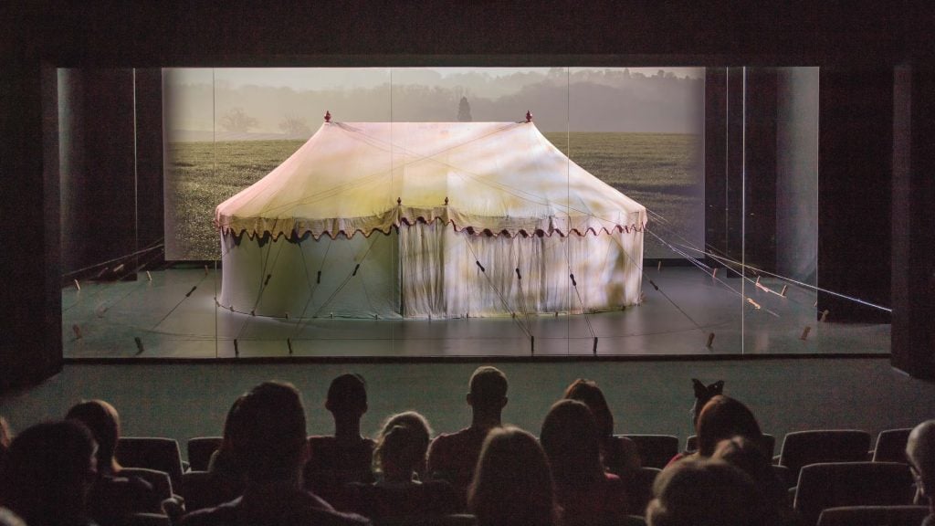 George Washington's Revolutionary War tent, white with scalloped red-trimmed edges, sits on a stage in a darkened room in front of an audience at the Museum of the American Revolution. Photo courtesy of the Museum of the American Revolution.