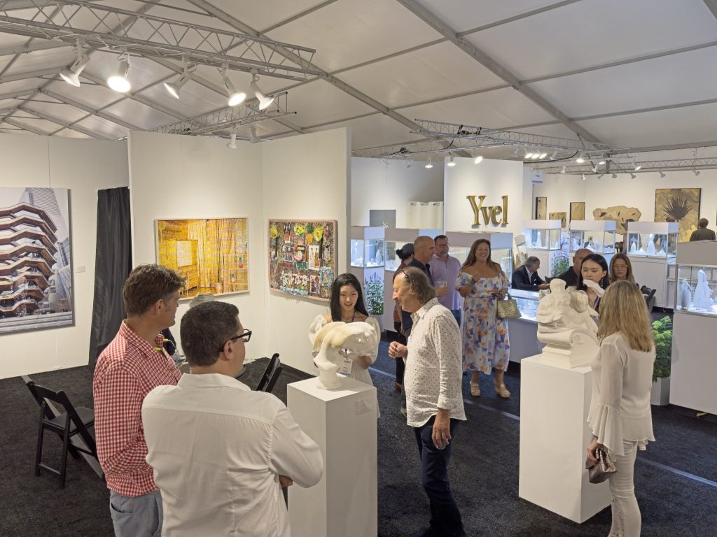 Inside the Hamptons Fine Art Fair tent with a few booths installed with art viewable and a number of visitors milling about.