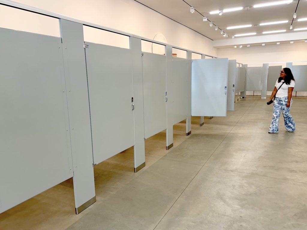 A woman in an art gallery looks at a line of grey bathroom stalls