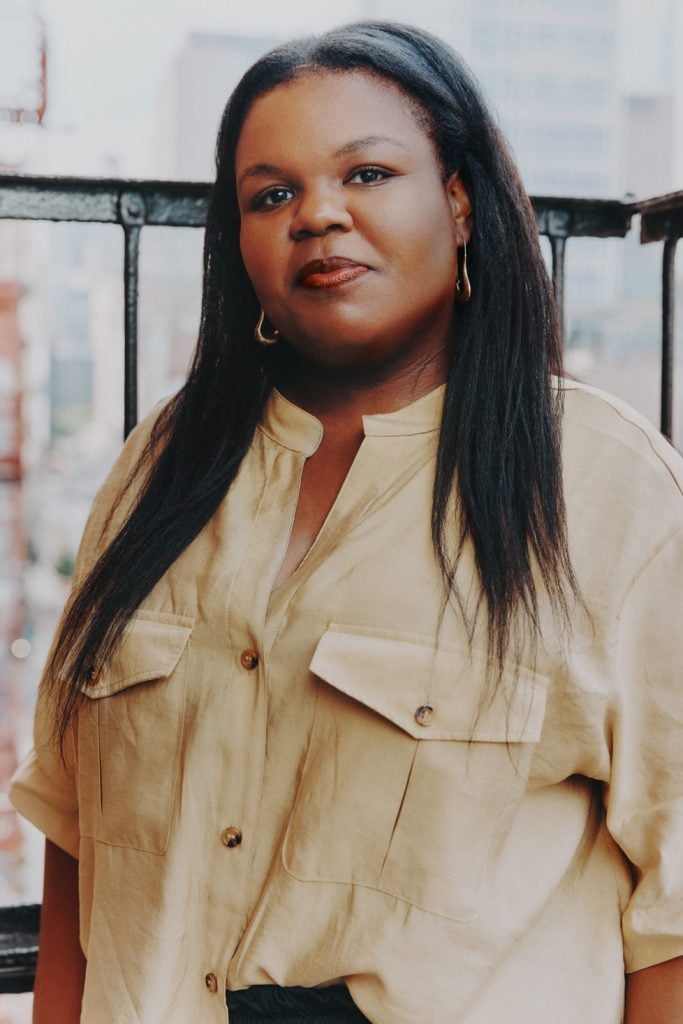 A color photograph shows a Black woman with long hair wearing a tan shirt.