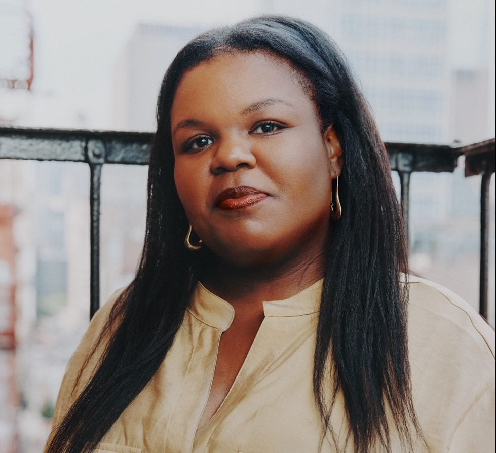 A color photograph shows a Black woman with long hair wearing a tan shirt.