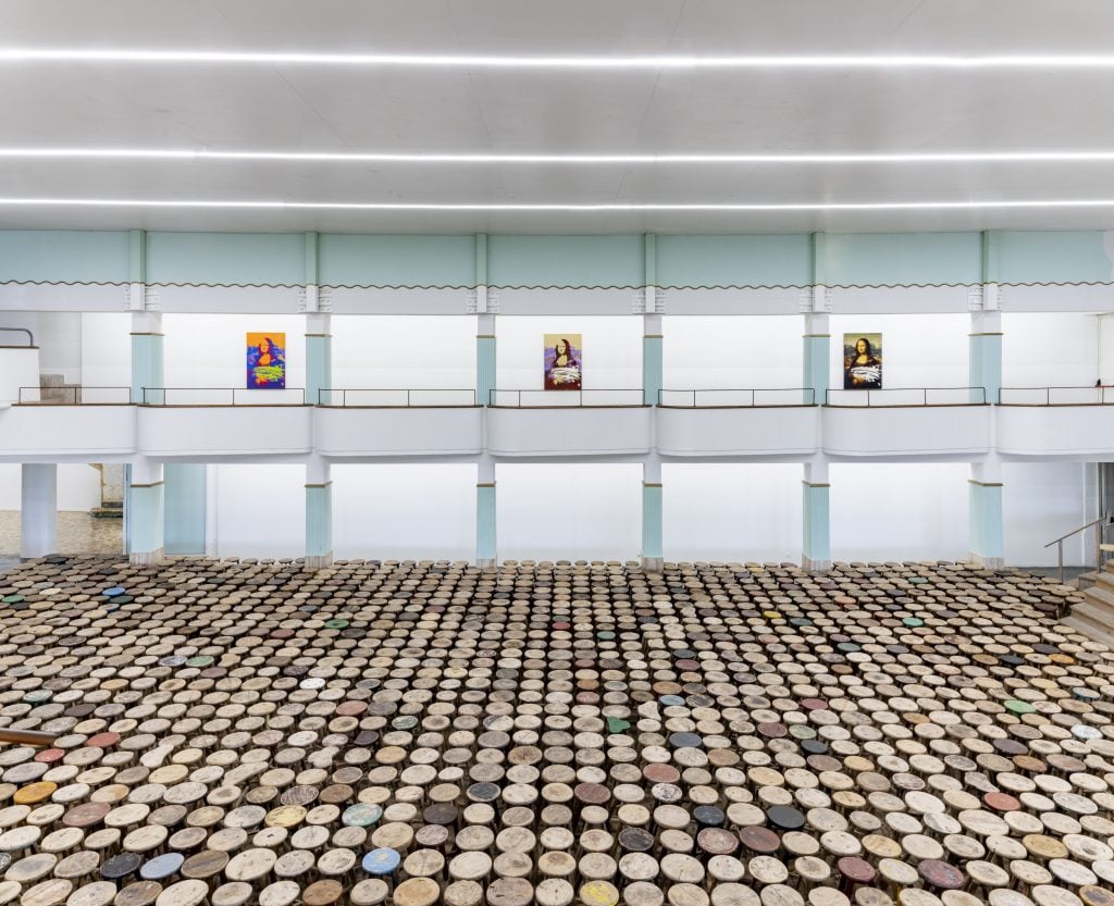 View across a large, two story gallery space with balcony, on the second story three variations on the mona lisa are installed, and across the entire floor hundreds of cylindrical objects are arranged in rows.
