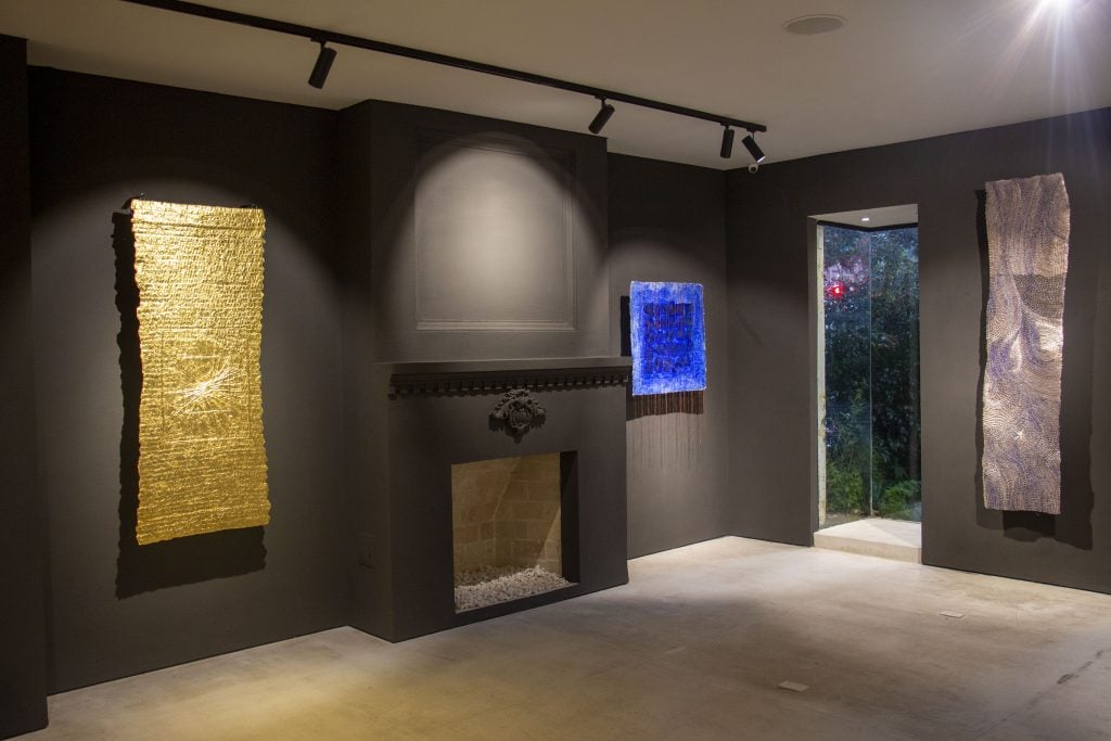 An interior room with dark grey walls and cream carpeting with a selection of textile works, including ones made of gold and lapis, hung on the walls by artist olga de amaral.