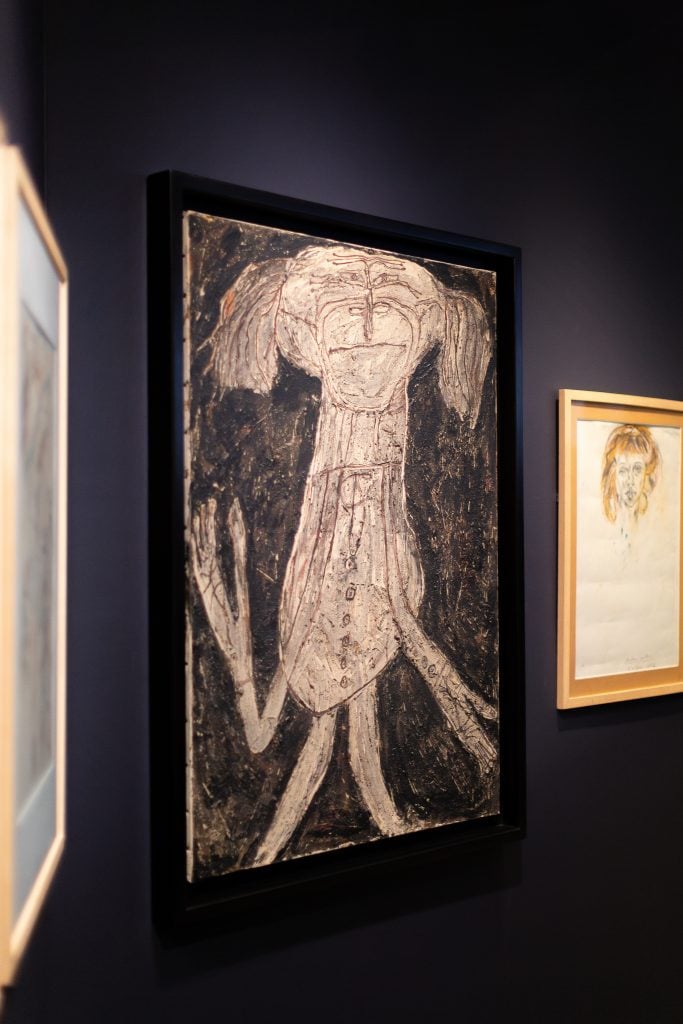 Inside the art brut exhibition at the American Folk Art Museum showing three artworks on a black wall, the center image a black and white figurative work by Jean Dubuffet.