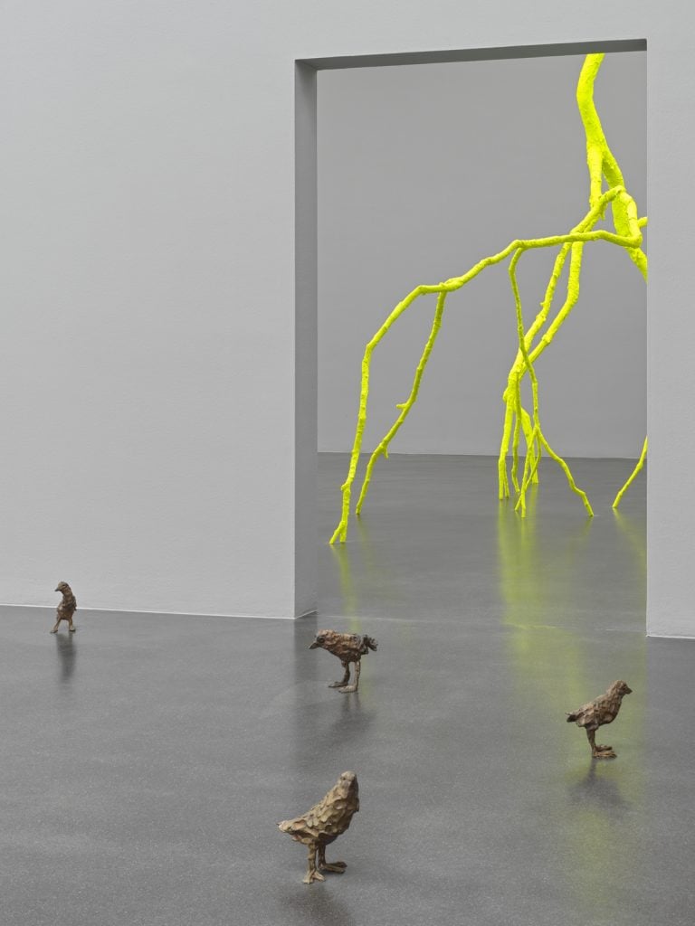 Installation view of "Ugo Rondinone: Cry Me a River" (2024) featuring primitive (2011–2012), comprised of four small-scale bird sculptures arranged on the floor, and viewable through a doorway lighting (2023), which is a florescent, large-scale sculpture that looks like a lightning strike. Walls and floor are both a muted grey.