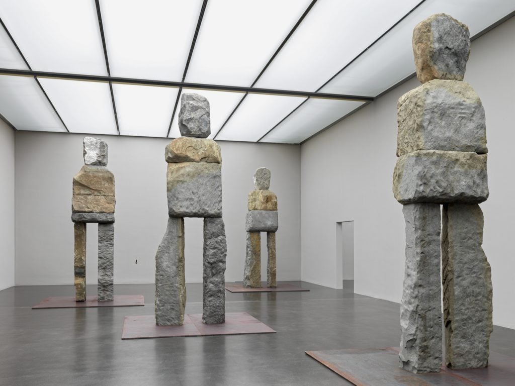 Four monumental sculptures in a grey gallery space made of stone that abstractly look like human figures.