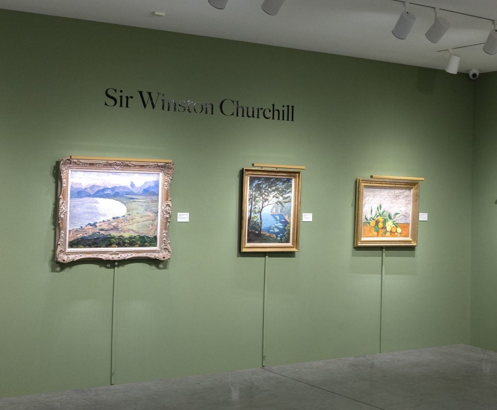 Installation view of entryway to Winston Churchill exhibition at Heather James Fine Art, with three landscape paintings in gold frames installed on a sage green wall that also has a vinyl graphic over top reading Sir Winston Churchill.