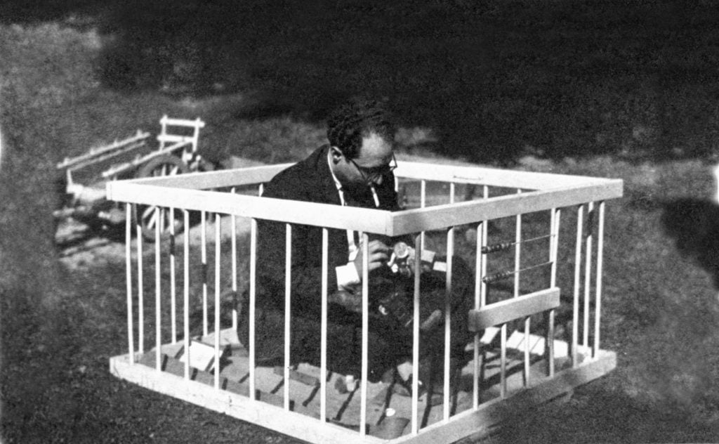 Black and white photograph of psychiatris Francesc Tosquelles sitting in what appears to be a childrens play pen with a cigarette hanging out of his mouth looking down at something.