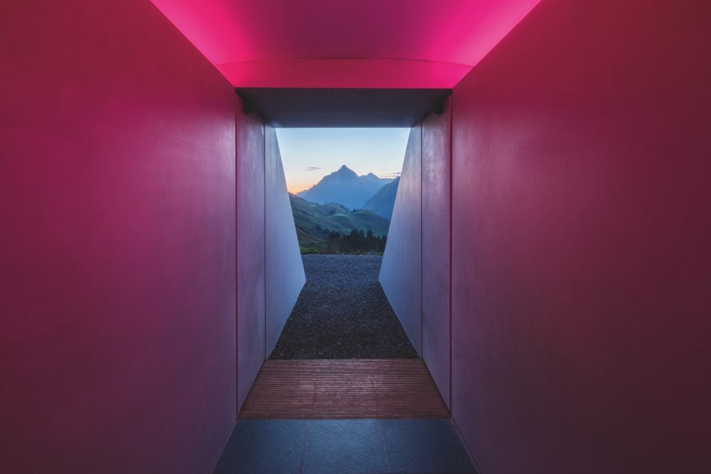 A view from inside a land art installation by James Turrell looking out a concrete tunnel filled with magenta light and the top of a mountain visible in the far distance.