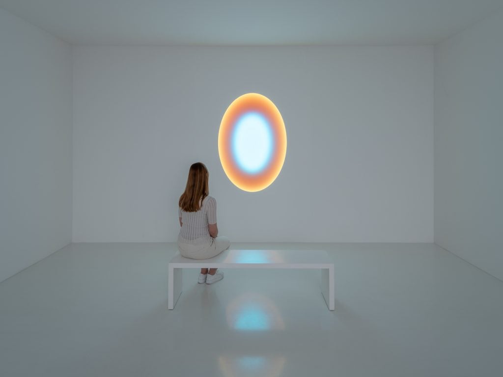 James Turrell installation of a small oval window of concentric colored light within a white cube gallery space with a white bench in front and a woman wearing all cream sitting on it.