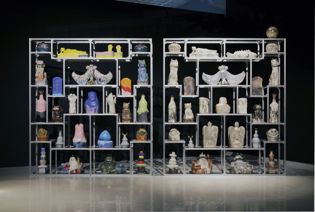 A metal shelving unit with various sculptural objects relating to artifacts and futuristic objects in a blackened out room, featured in the exhibition I Am Because We Are.