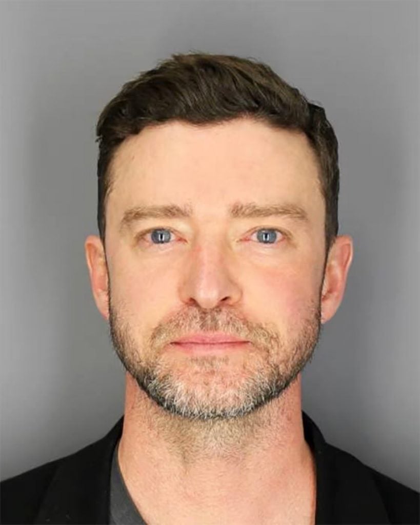 Justin Timberlake stares straight ahead at the camera in a standard mugshot. He has a light beard and is wearing a black t-shirt. 