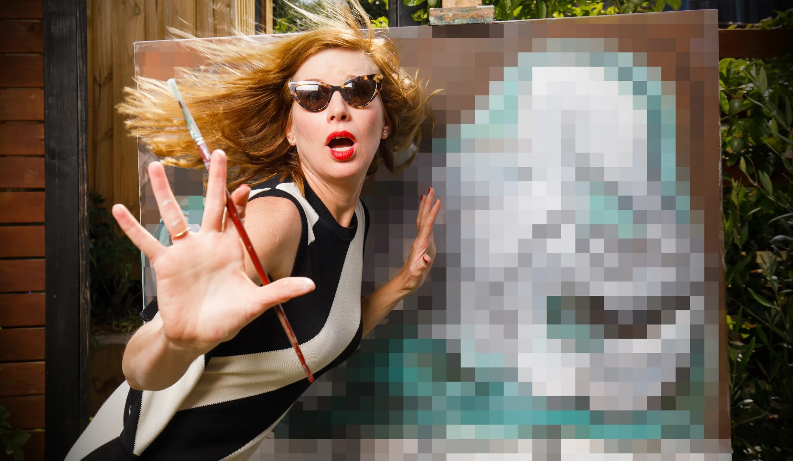 Artist Kirsha Kaechele in sunglasses poses with a paintbrush in front of an artwork.