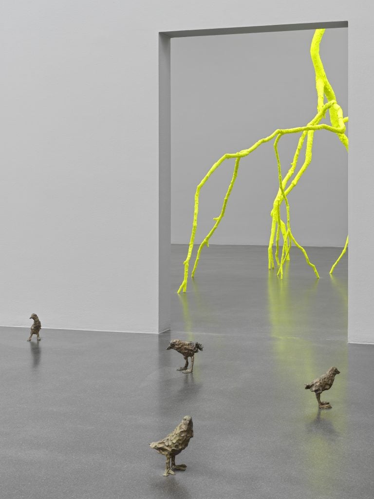small bronze bird sculptures on the floor of a gallery, a fluorescent lightning sculpture is visible behind them