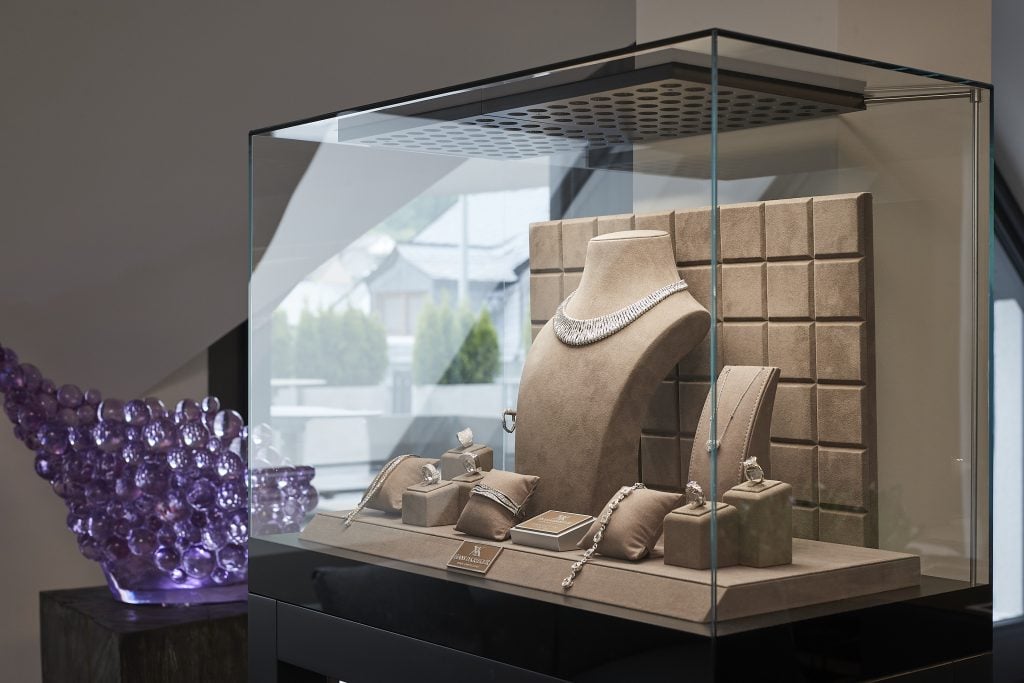 A glass vitrine showcasing a range of different types of diamond jewelry with a purple glass sculpture by Cedric and Pierre Koukjian that looks like grapes viewable just behind, both as part of the koukjian x krieger collab.