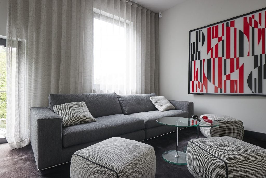 Inside the space created by Andreas Krieger for koukjian x krieger with a grey sofa, two cloth ottomans, round glass nesting coffee table, and an abstract black, white, and red artwork on the wall.