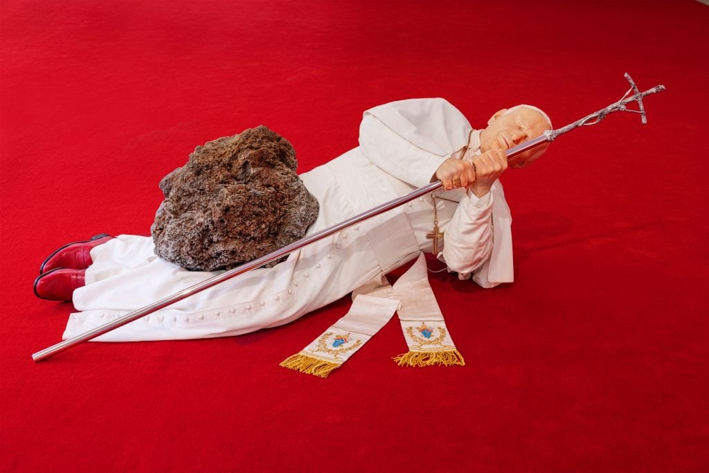 a model of a pope is lying on the floor clutching a long gold papal pole as though it might help me get up even though it won't