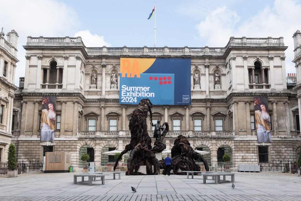 a large installation that looks like sprawling roots of a tree fills the central court of a grand but grey old fashioned building that has a sign hanging on it that says 