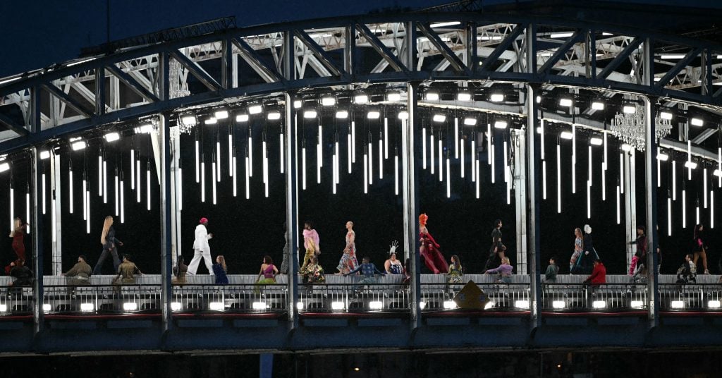 Models walk along a catwalk on the Passerelle Debilly bridge, illuminated at night, during the Olympic opening ceremonies in Paris. The scene's resemblance to the Last Supper sparked controversy among conservative Christians.