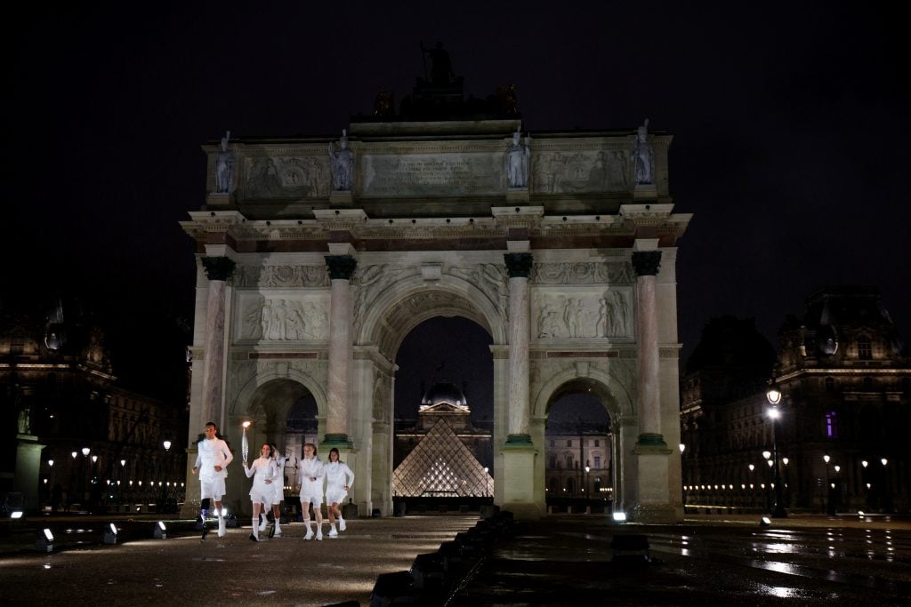 Five runners, one of them bearing the Olympic torch, running away from the Louvre Museum