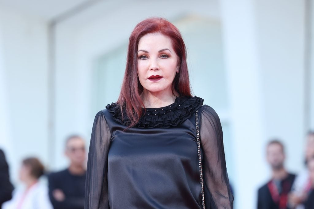 Priscilla Presley at a red carpet event in a sleek black top. She has straight dark red hair to her shoulders in a side part, and matching dark red lipstick. 