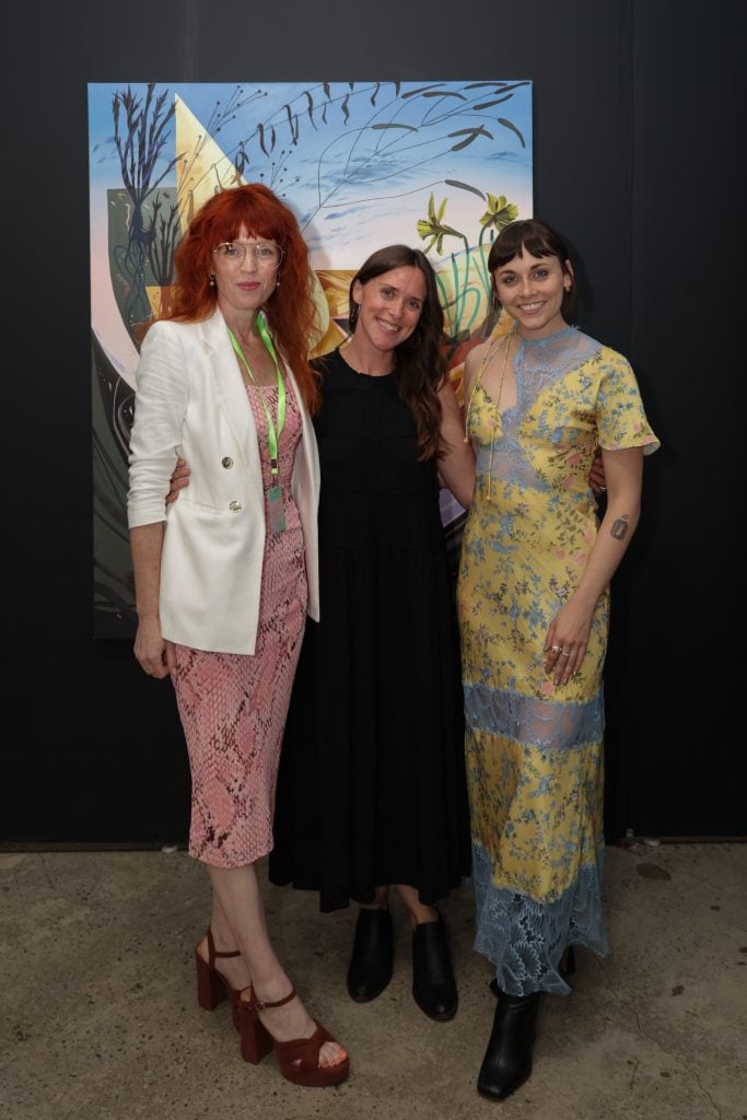 Three well-dressed women stand in front of a painting