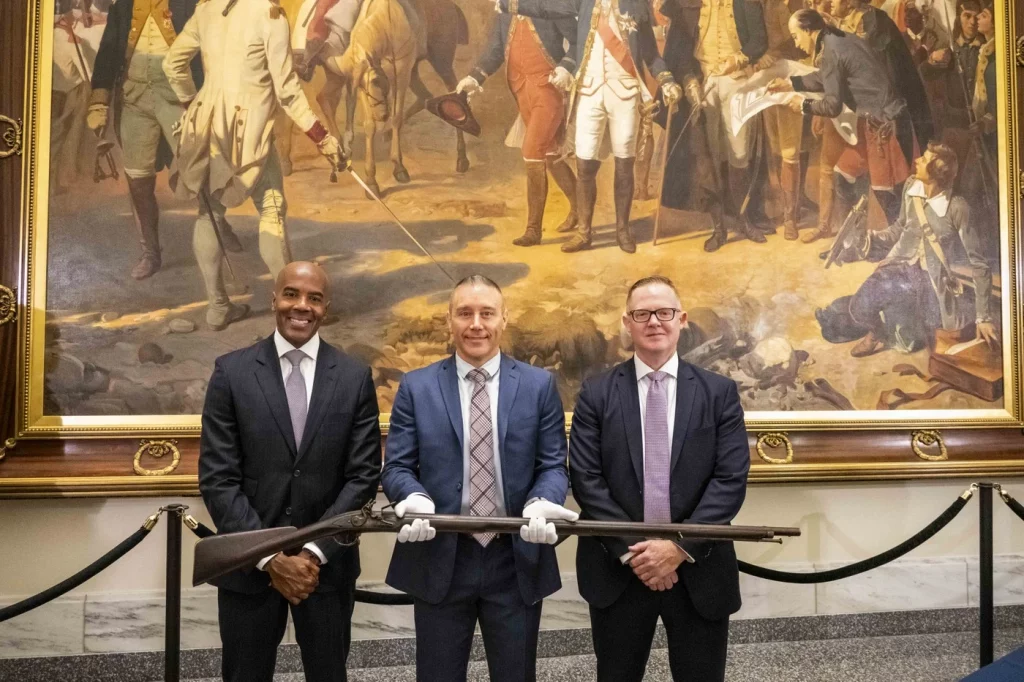 Three FBI agents hold a stolen Revolutionary War musket in a ceremony returning it to the Museum of the American Revolution. They stand in front of a history painting of the Revolutionary War titled 
