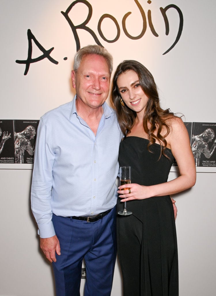 Robert Bowman wearing a light blue button down and navy trousers standing next to his daughter Mica Bowman wearing a black strapless dress holding a champagne flue in front of the intro wall of the Rodin exhibition at Bowman Sculpture.