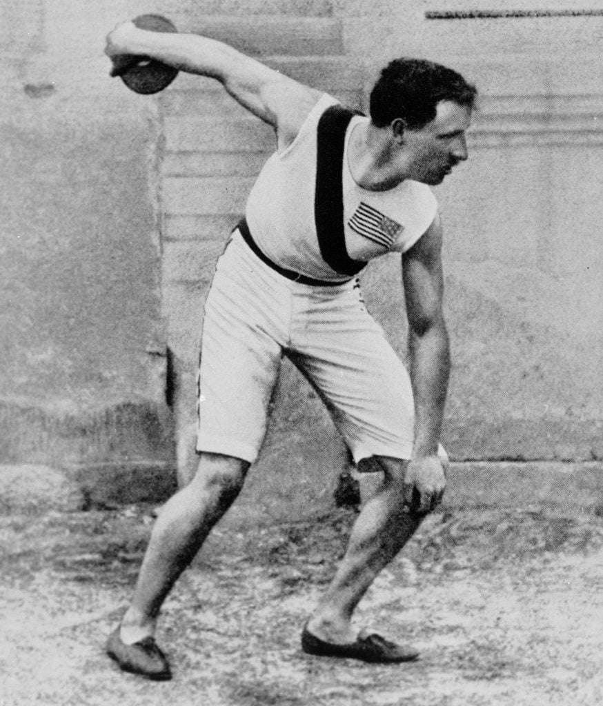 Black and white photo of Robert Garrett standing in position to throw a discus as part of the first modern olympics, wearing white shorts and a white tank with an american flag on his chest.