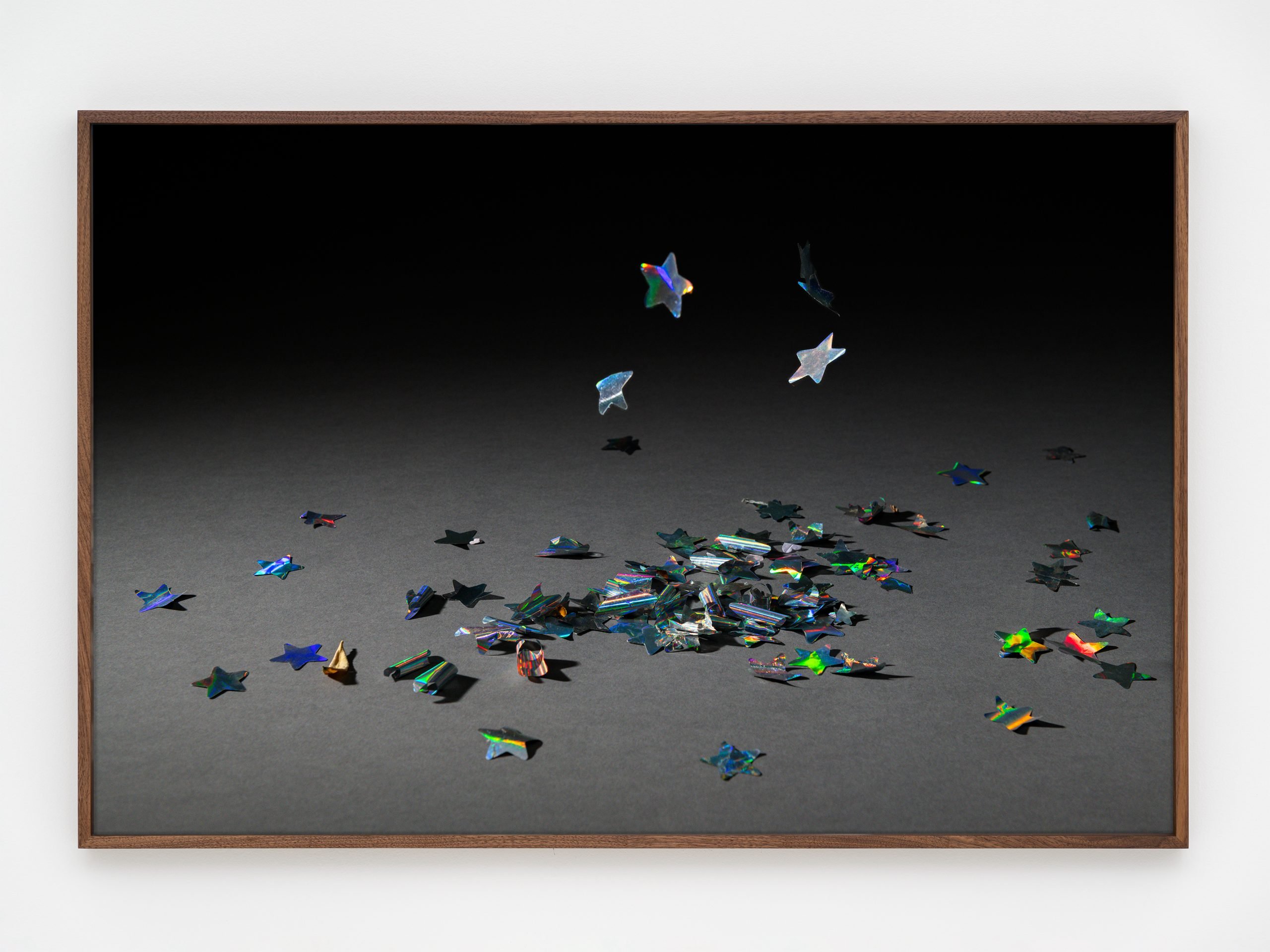 A photo by rose salane of a small pile of silver star shaped confetti on a grey ground and diffuse black background.