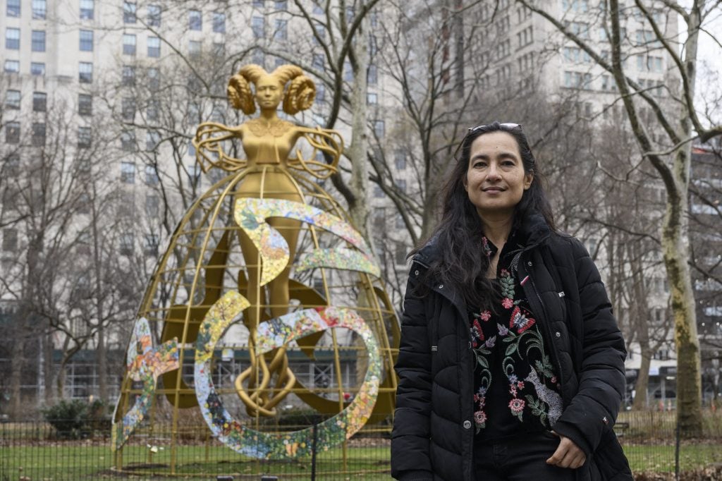 Artist Shahzia Sikander standing in front of her gold sculpture of a woman with circular braids and roots for arms and legs