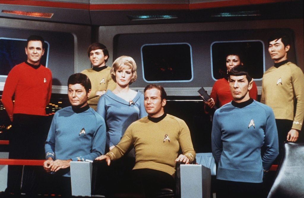 The cast of Star Trek, dressed in colorful uniforms, gathered around the deck of a spaceship set