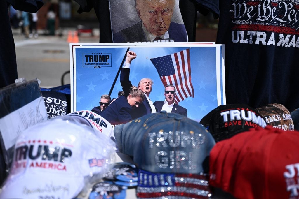 An image of a man raising his fist in front of an American flag on a table full of hats that say TRUMP