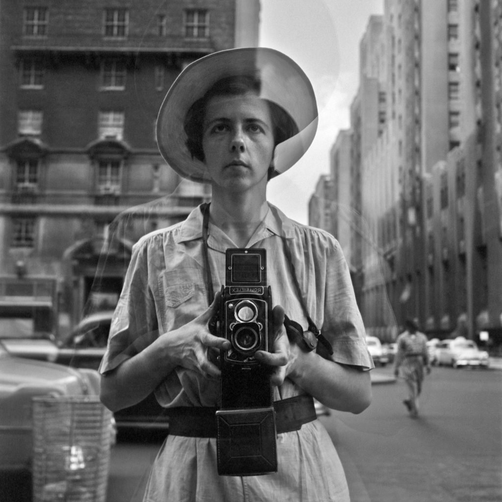 Black-and-white self portrait of street photographer vivian maier taken in a window reflection on a new york city street in the 1950s.