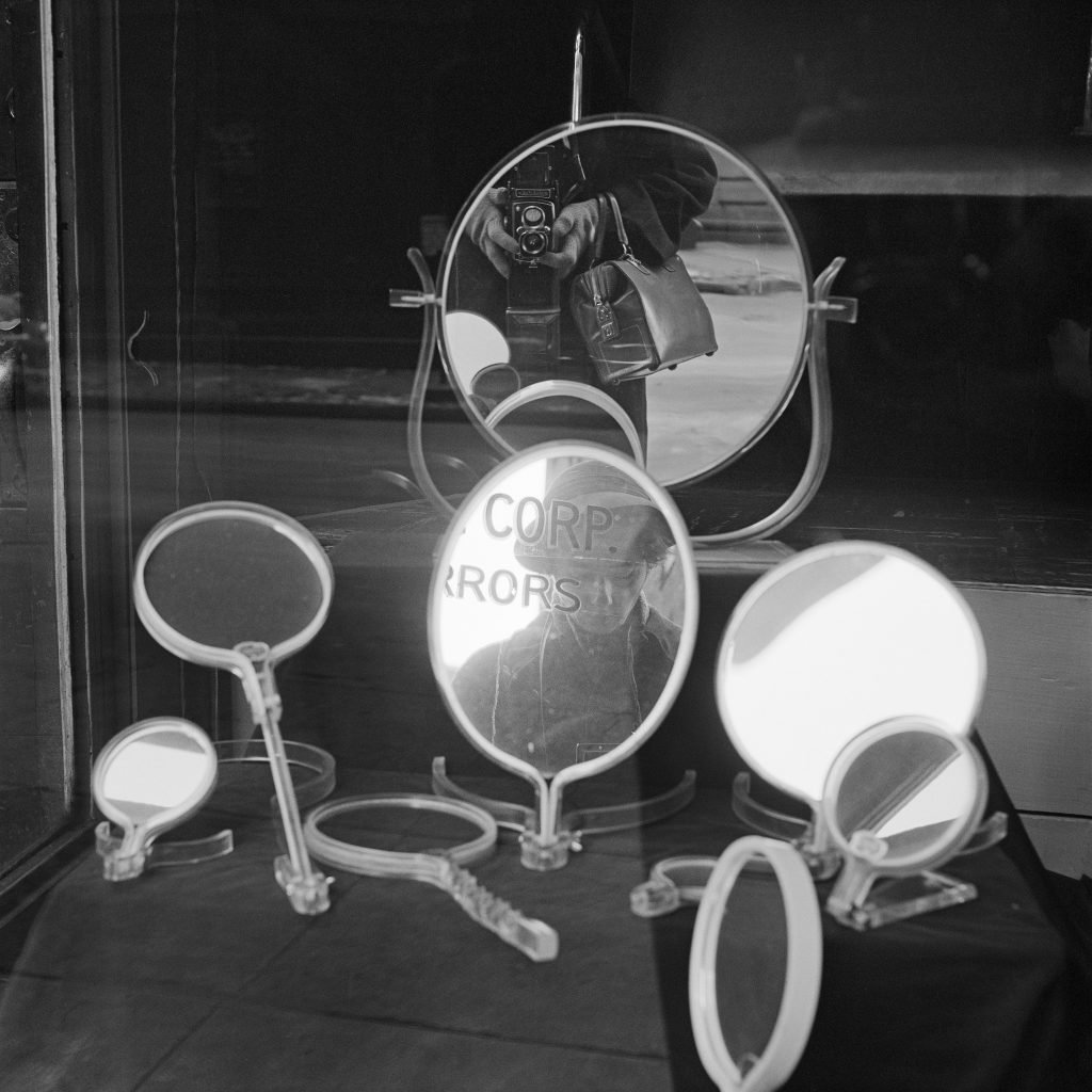 Black-and-white portrait taken by street photographer Vivian Maier with the artists reflection and camera reflection seen in a window display of handheld and tabletop mirrors.