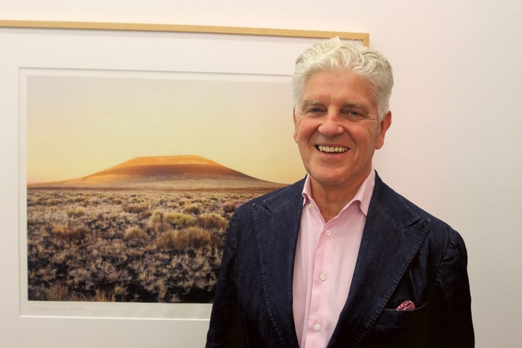 Wolfgang Häusler standing in front a photo of a land art project by James Turrell wearing a pink button down shirt and black suit jacket.