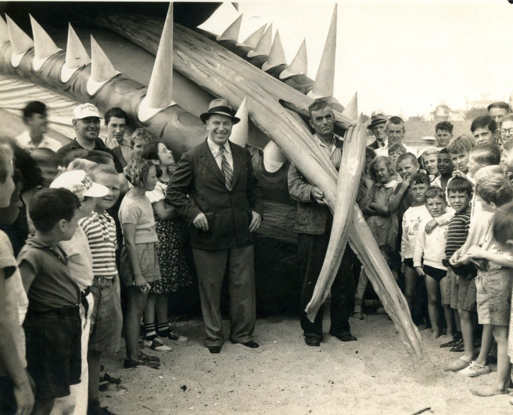 Tony Sarg stands in a black and white photo in front of his sea monster surrounding by people