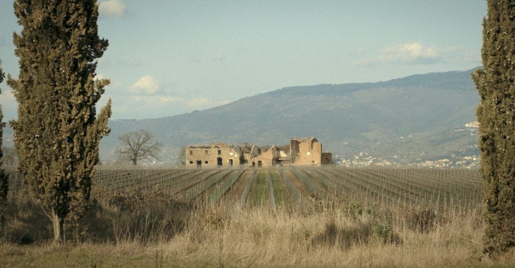 an ancient tuscan stable is in the distance of rolling fields with mountains behind it
