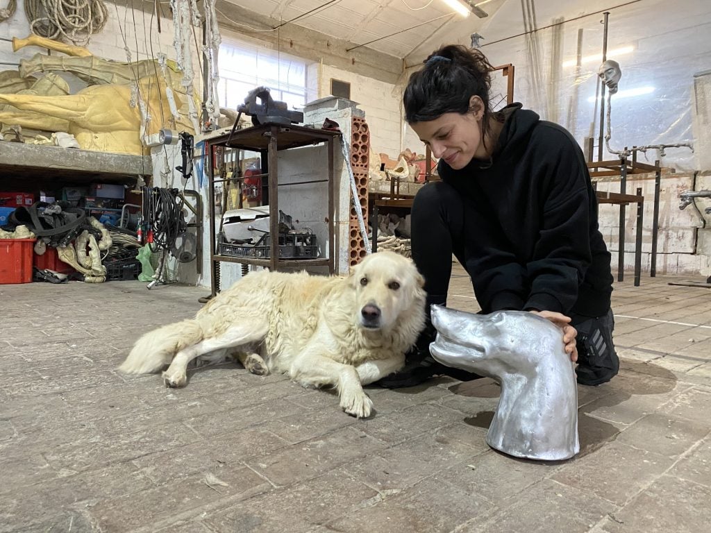 a woman posed in her artist's studio next to a white dog a sculpture she made of the animal