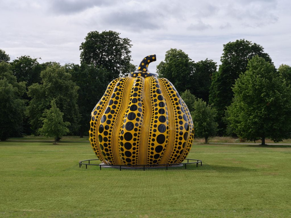 a large pumpkin is instaleld in a green space with trees in the background, it has vertical stripes of black dots of various sizes 