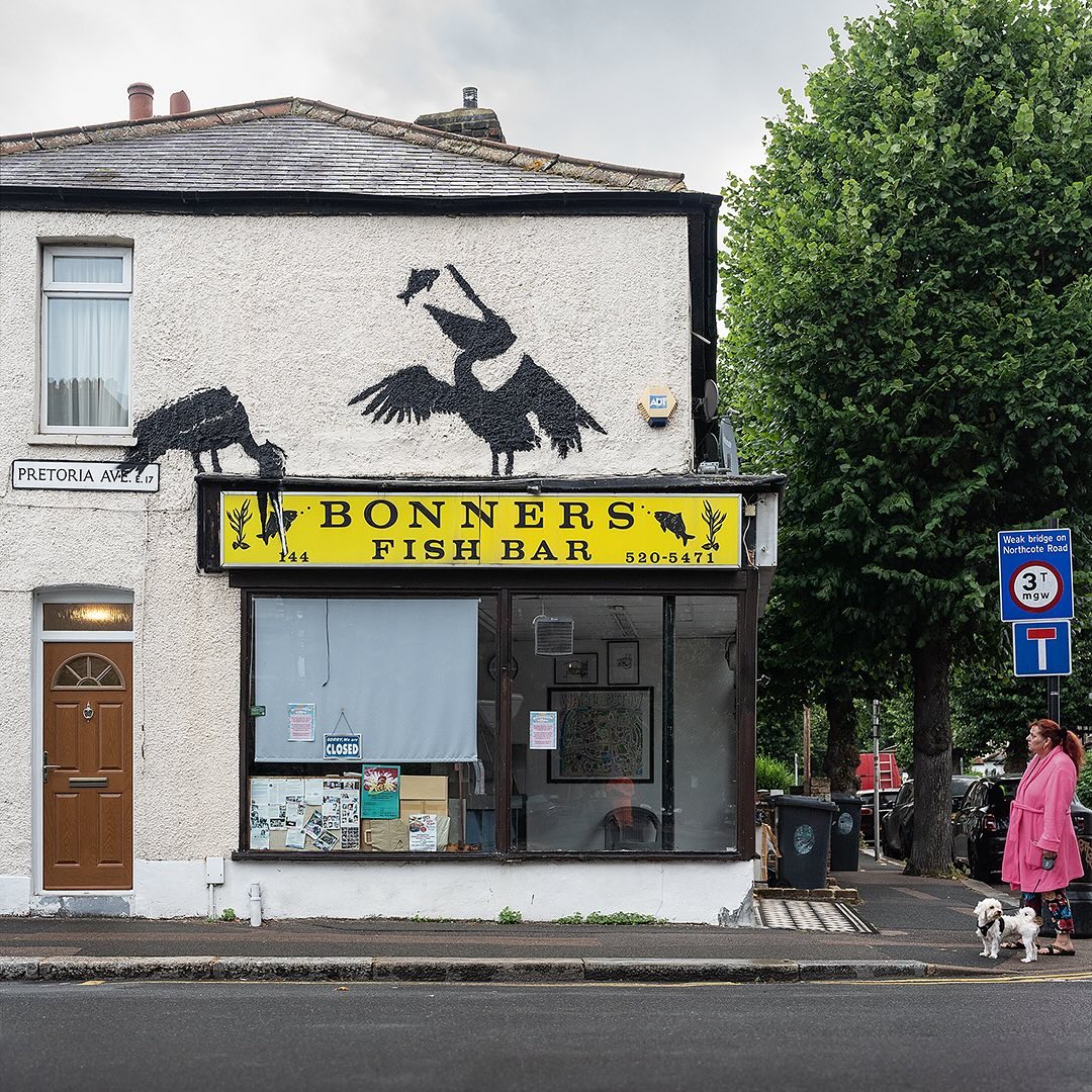 Banksy stenciled the silhouette of a pair of pelicans perched above the yellow sign of Bonners Fish Bar, one appearing to eat a fish off the sign. A woman in a pink dressing gown walking a small white dog stands facing the building on the corner.