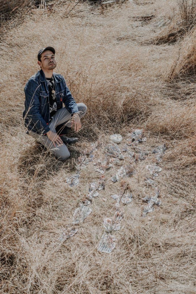 Alejandro Garcia Cortreras, wearing a baseball hat, a dark denim jacket, and gray pants, kneels next to some of his small ceramic sculptures, laid out in the dry brush of the landscape like ancient bones being unearthed in an archaeological dig.