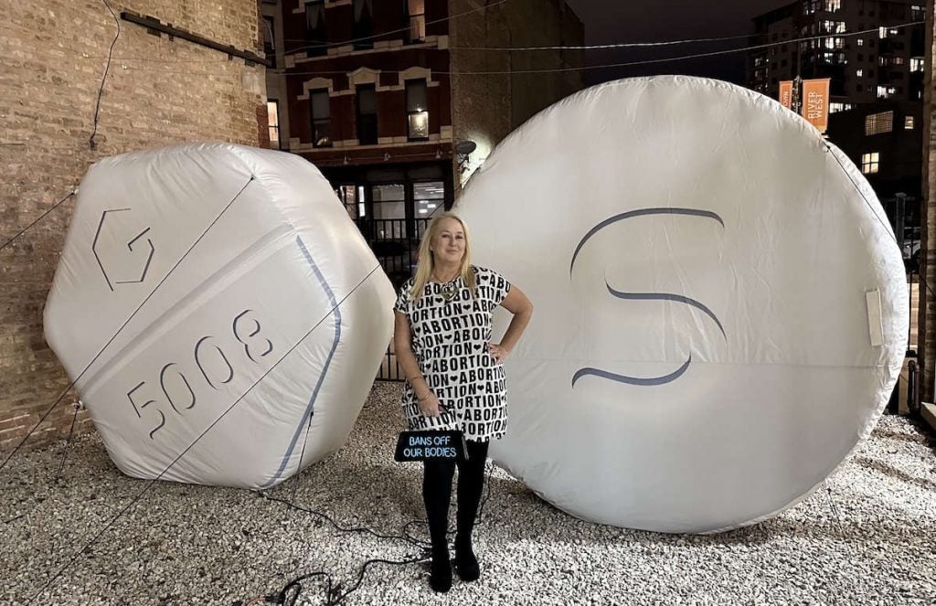 Michele Pred und ihre aufblasbare Skulptur <em>Abortion is Healthcare (Mifepristone and Misoprostol)</em>. Wearing a white dress printed with the word ” vote ” in black block lettering “, the artist holds a clutch purse with light up message reading off our bodies. she stands front of the two towering sculptures blown versions of medical abortion pills. width=”1024″ height=”663″ srcset=”https://news.artnet.com/app/news-upload/2024/08/Abortion-is-Healthcare_Michele-Pred-1024×663.jpg 1024w, https://news.artnet.com/app/news-upload/2024/08/Abortion-is-Healthcare_Michele-Pred-300×194.jpg 300w, https://news.artnet.com/app/news-upload/2024/08/Abortion-is-Healthcare_Michele-Pred-50×32.jpg 50w, https://news.artnet.com/app/news-upload/2024/08/Abortion-is-Healthcare_Michele-Pred.jpg 1200w” sizes=”(max-width: 1024px) 0vw, 1024px”/></p>
<p id=